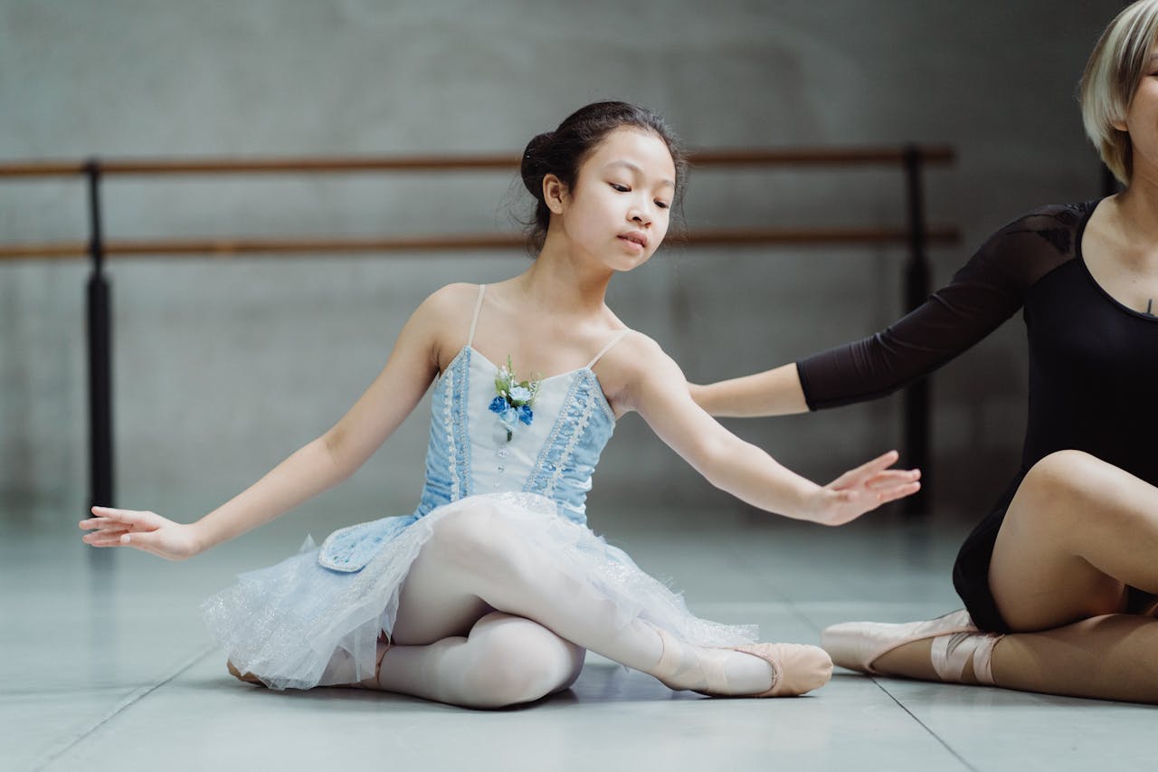 Full body of Asian girl sitting with legs crossed on floor with crop personal instructor in ballet studio