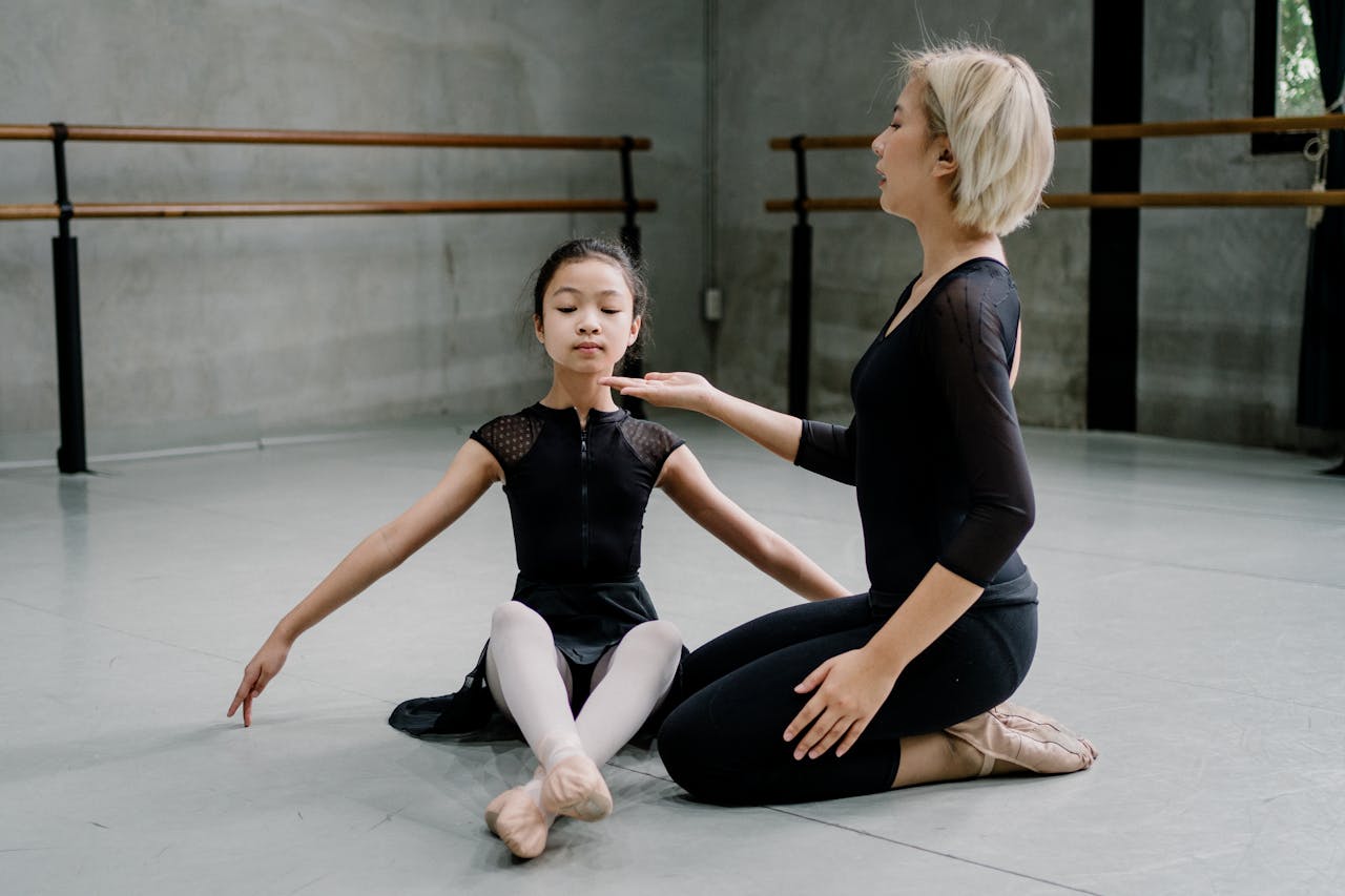 A dance instructor guides a young girl in a ballet class setting. Indoor studio environment.
