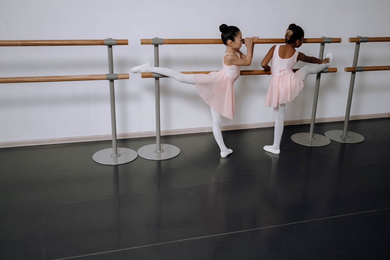 Two young ballerinas in pink tutus stretching at the barre in a ballet studio.